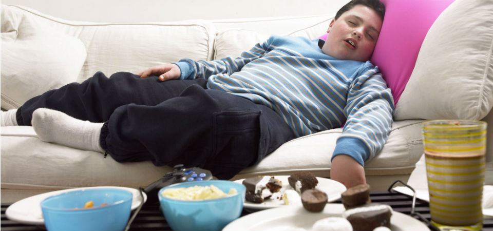Overweight boy sleeping on a sofa with a lot of unk food on table in his front.