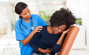image of a dark skinned woman in the hospital, clutching stomach in pain witha dark skinned female health professional beside her